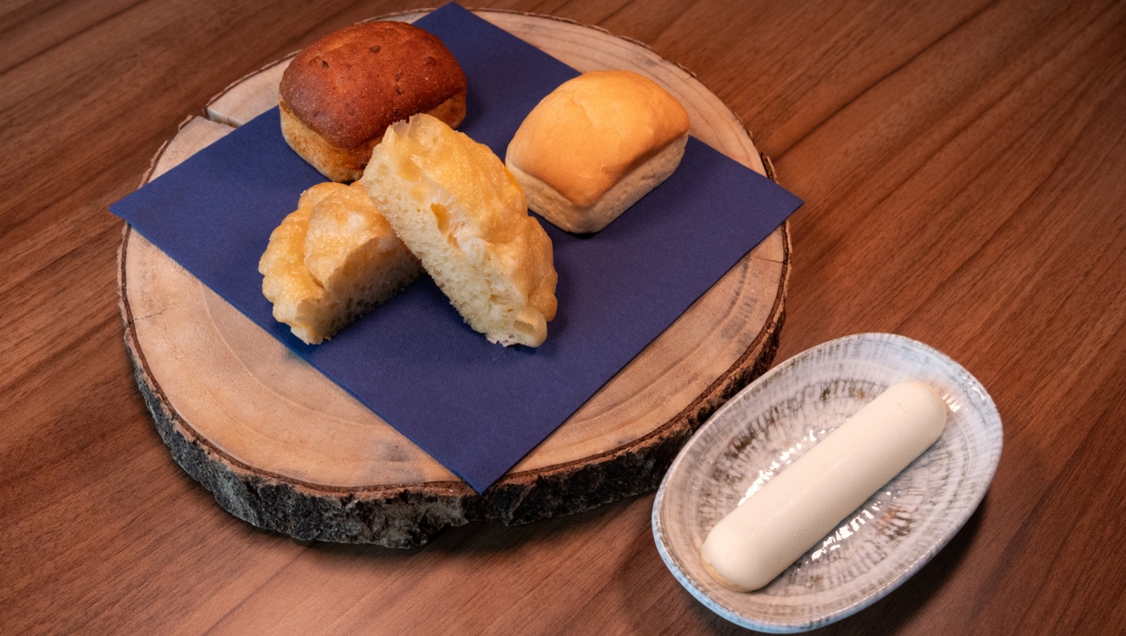 Trzy rodzaje pieczywa na drewnianej desce z niebieską serwetką oraz pałeczka masła na ceramicznym talerzu