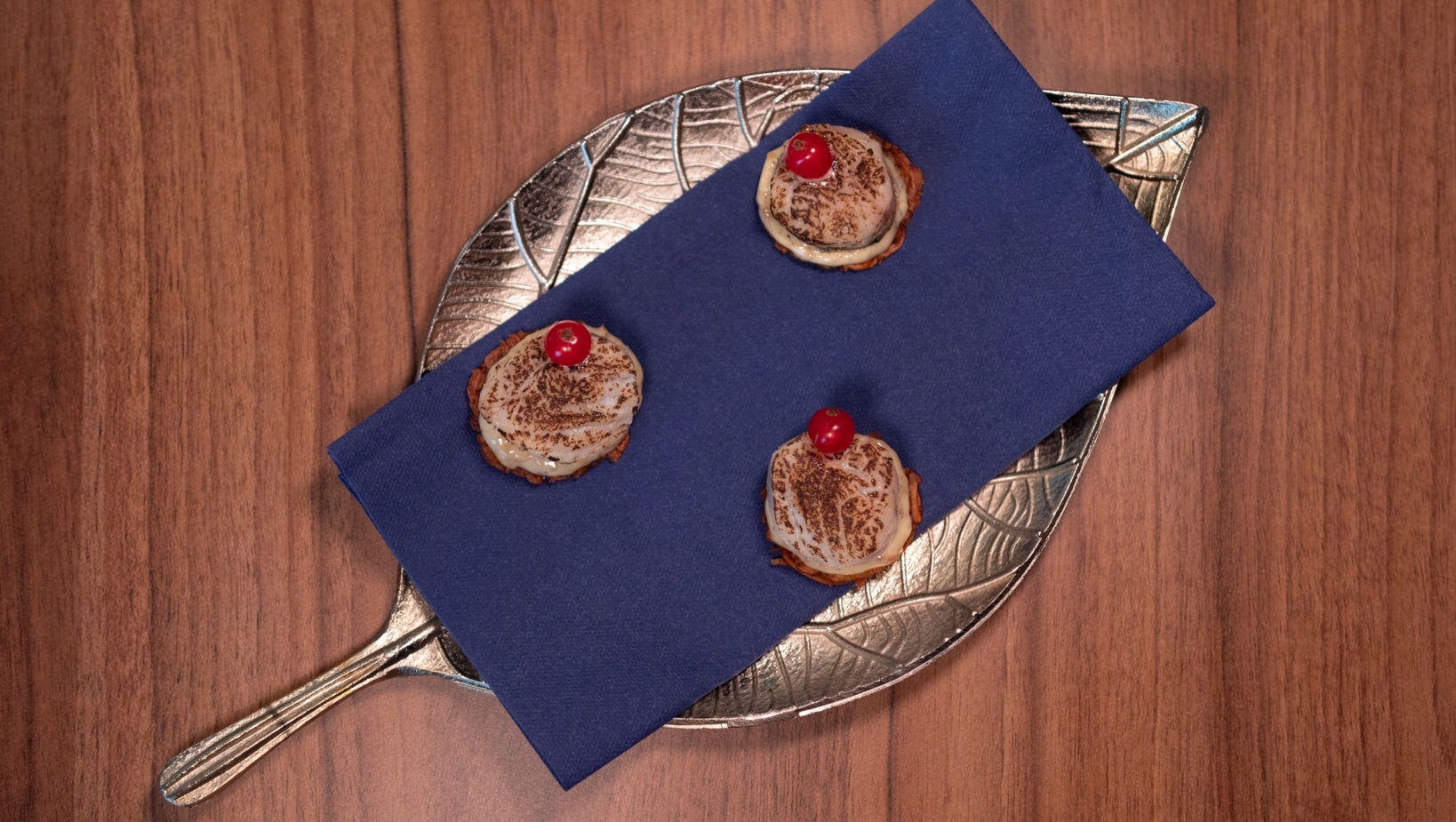 Trzy kremowe mini deserki z opalonym wierzchem i czerwonym owocem na srebrnej tacy z niebieską serwetką