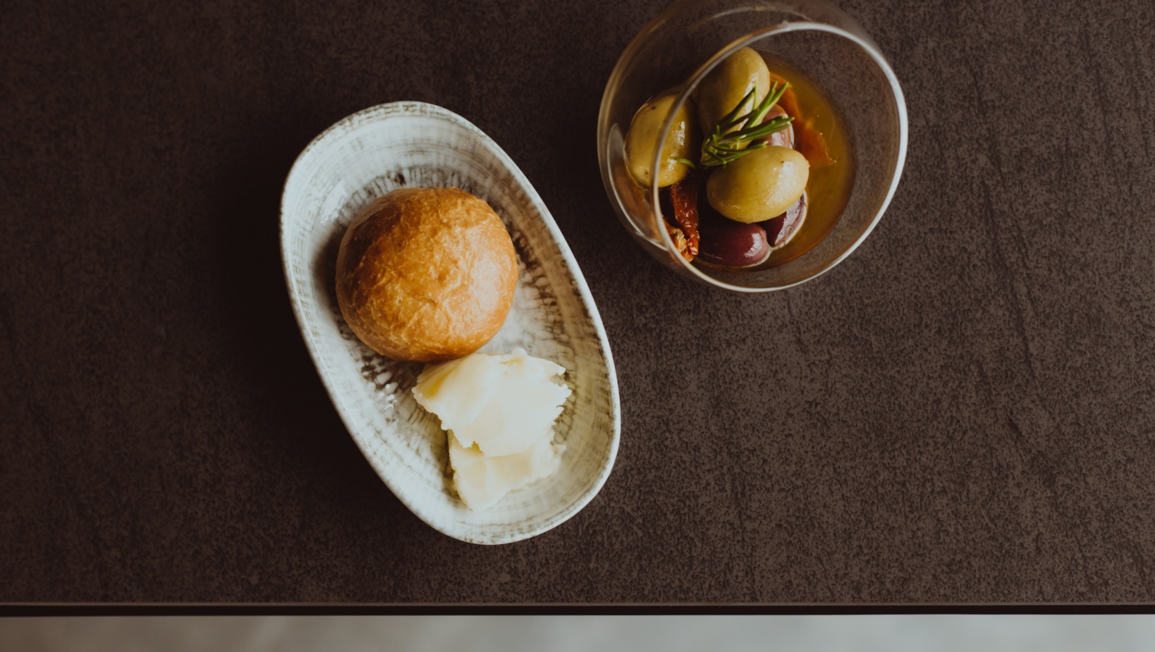 Mała miska z zielonymi i czarnymi oliwkami z rozmarynem oraz talerz z bułką i masłem na ciemnym stole