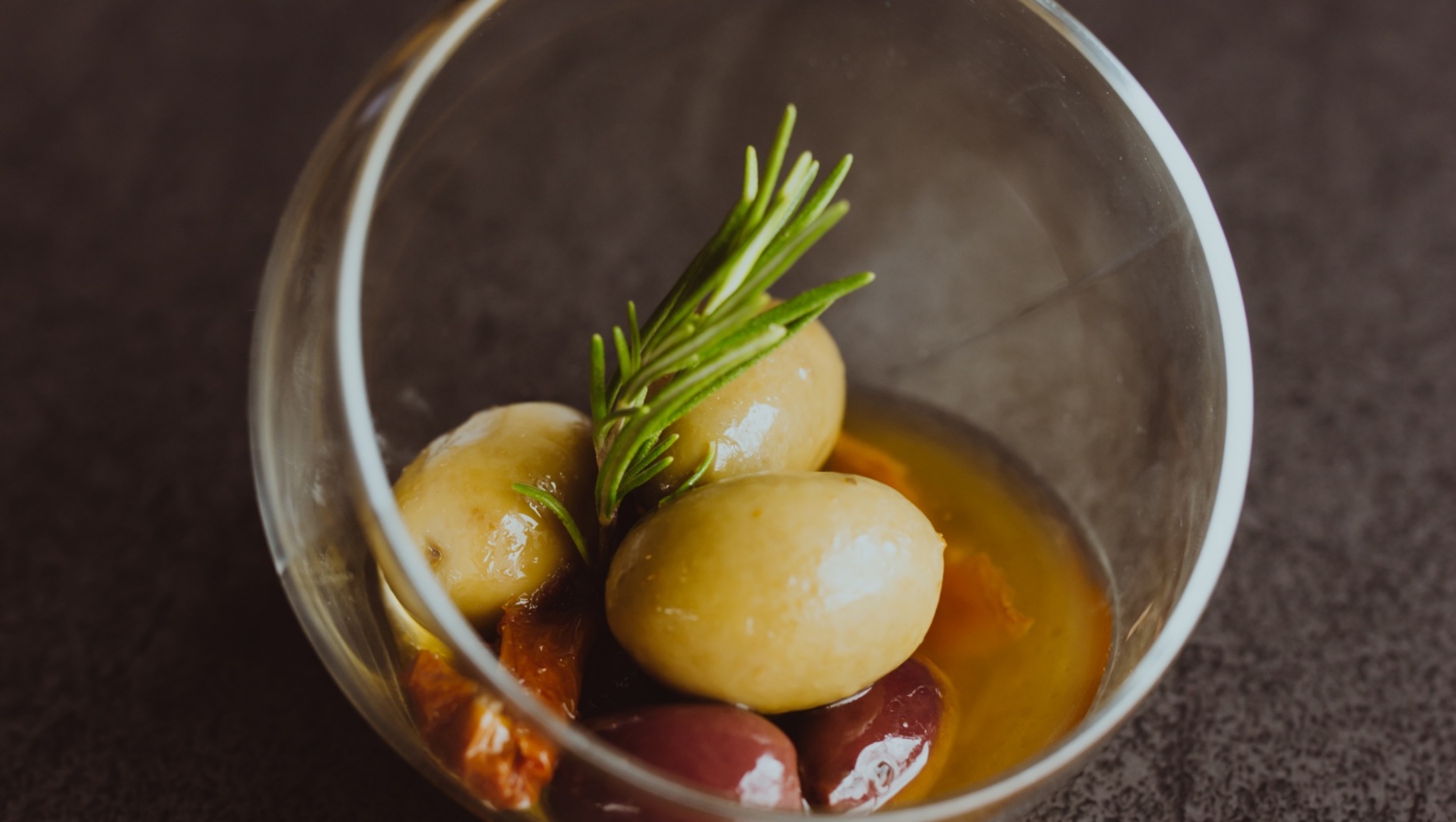 Białe i fioletowe oliwki w oliwie z oliwek z gałązką świeżego rozmarynu i suszonymi pomidorami w szklanej miseczce