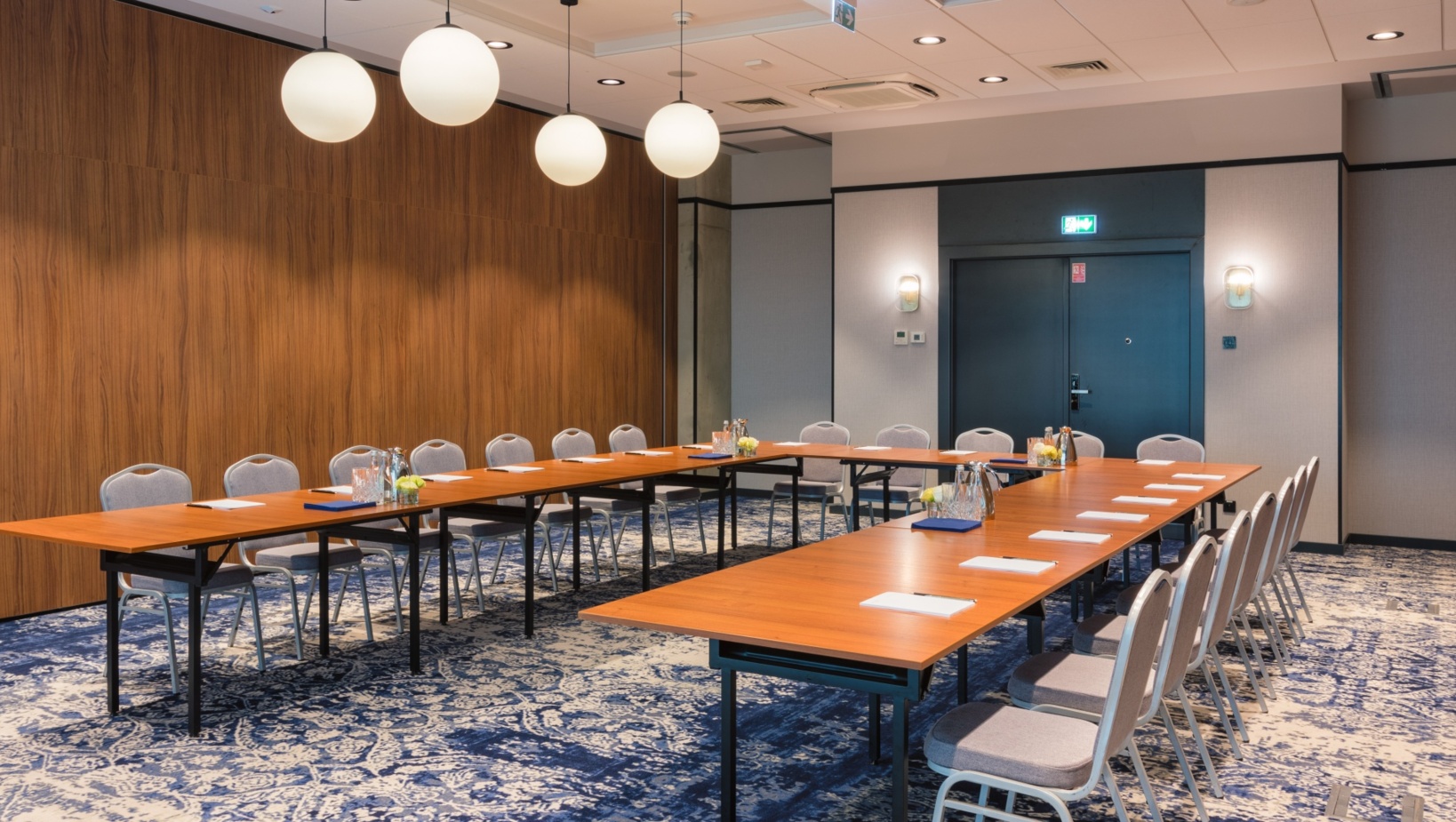 Sala konferencyjna z układem stołów w kształcie litery U, wyposażona w wygodne krzesła i nowoczesne oświetlenie kuliste.