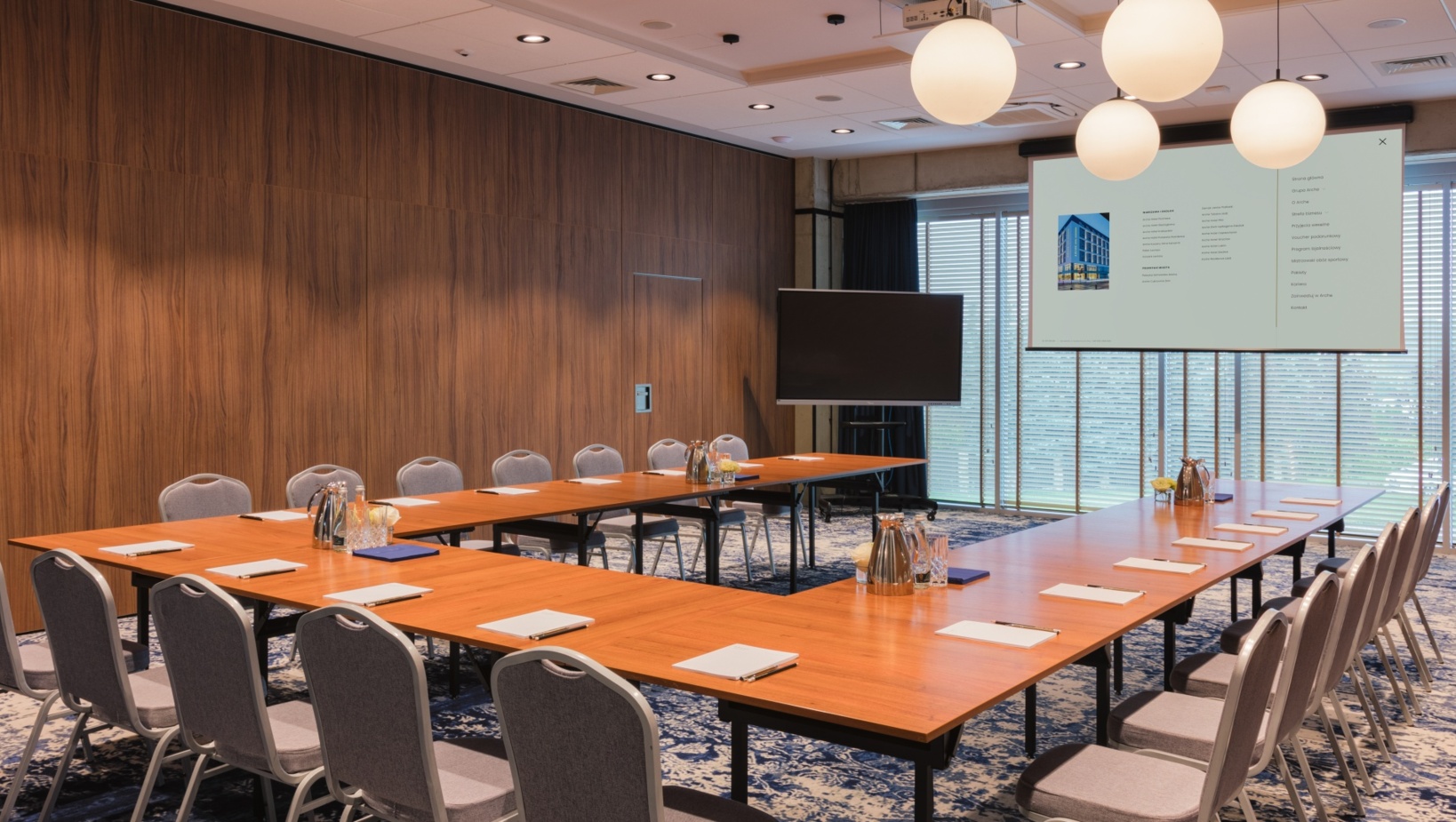 Sala konferencyjna z układem stołów w kształcie litery U, wyposażona w projektor i telewizor, wystrój drewniane panele i nowoczesne oświetlenie.