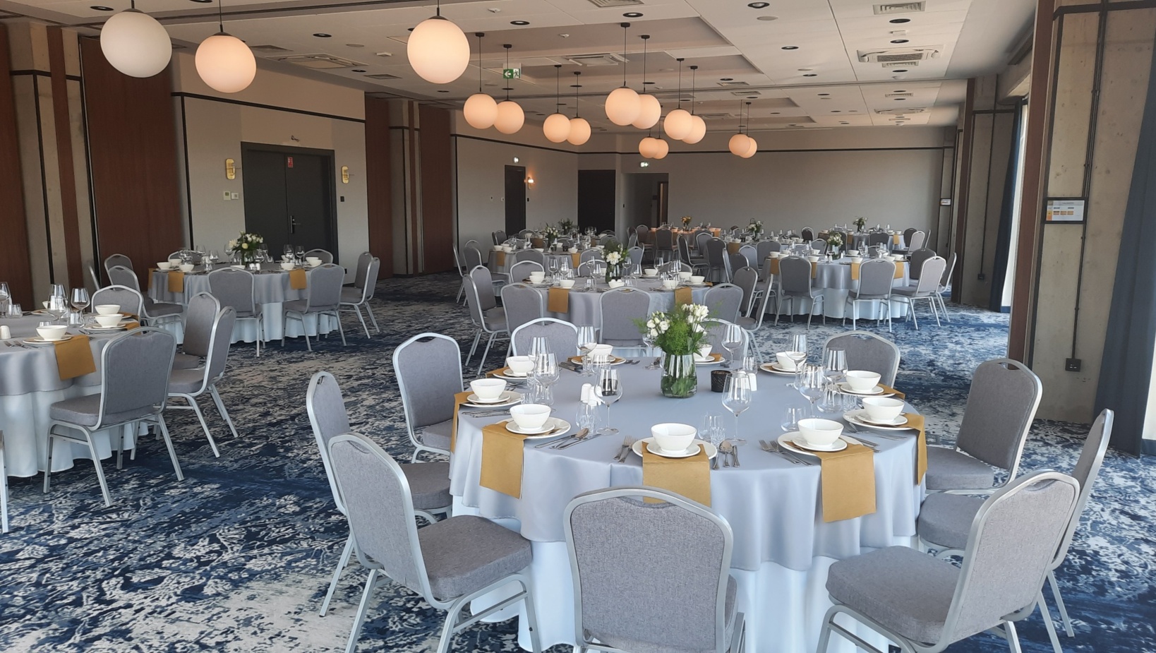 Sala bankietowa z okrągłymi stołami nakrytymi jasnoszarymi obrusami i żółtymi serwetkami, nowoczesne oświetlenie kuliste, kwiatowe aranżacje.