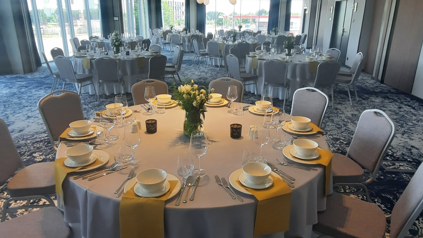 Sala bankietowa z okrągłymi stołami udekorowanymi białymi obrusem, żółtymi serwetkami i kwiatowymi aranżacjami