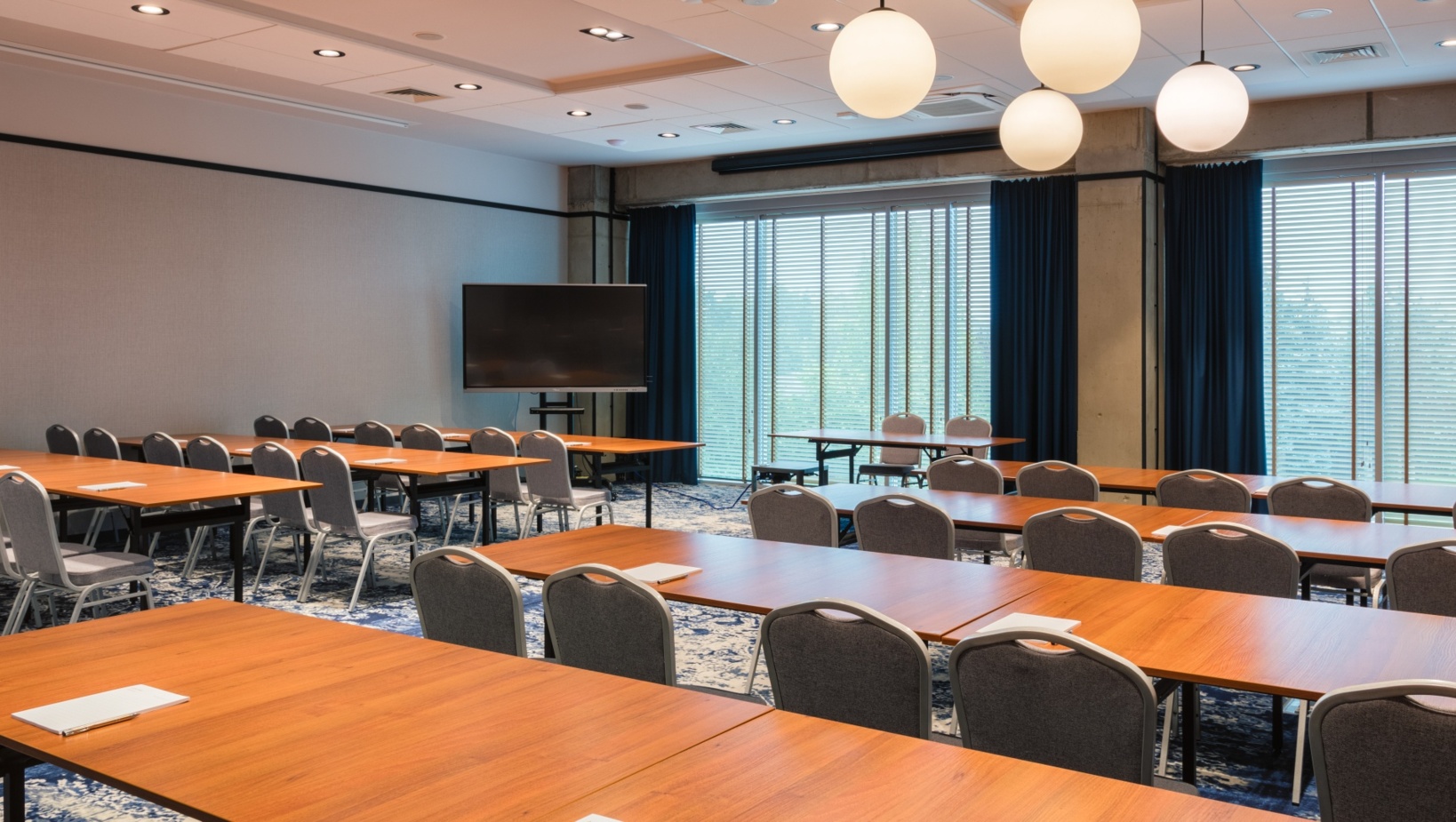 Przestronna sala konferencyjna z drewnianymi stołami, szarymi krzesłami i dużym ekranem do prezentacji przy świetle kulistych lamp.