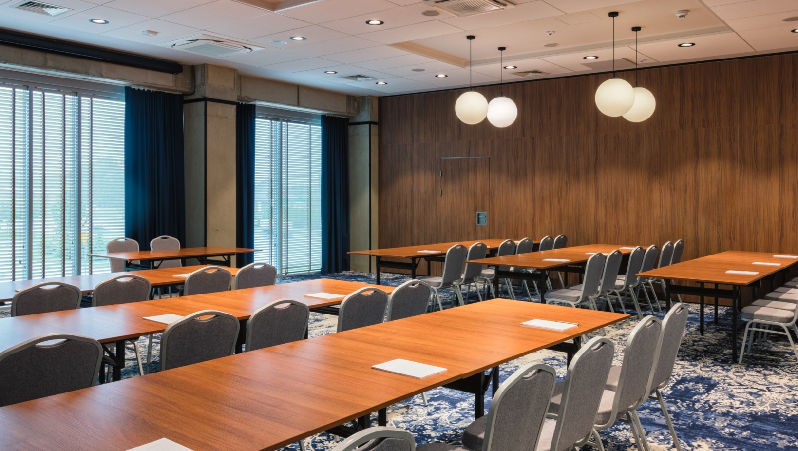 Nowoczesna sala konferencyjna z drewnianymi stołami, szarymi krzesłami, dużymi oknami i miękkim niebiesko-białym dywanem