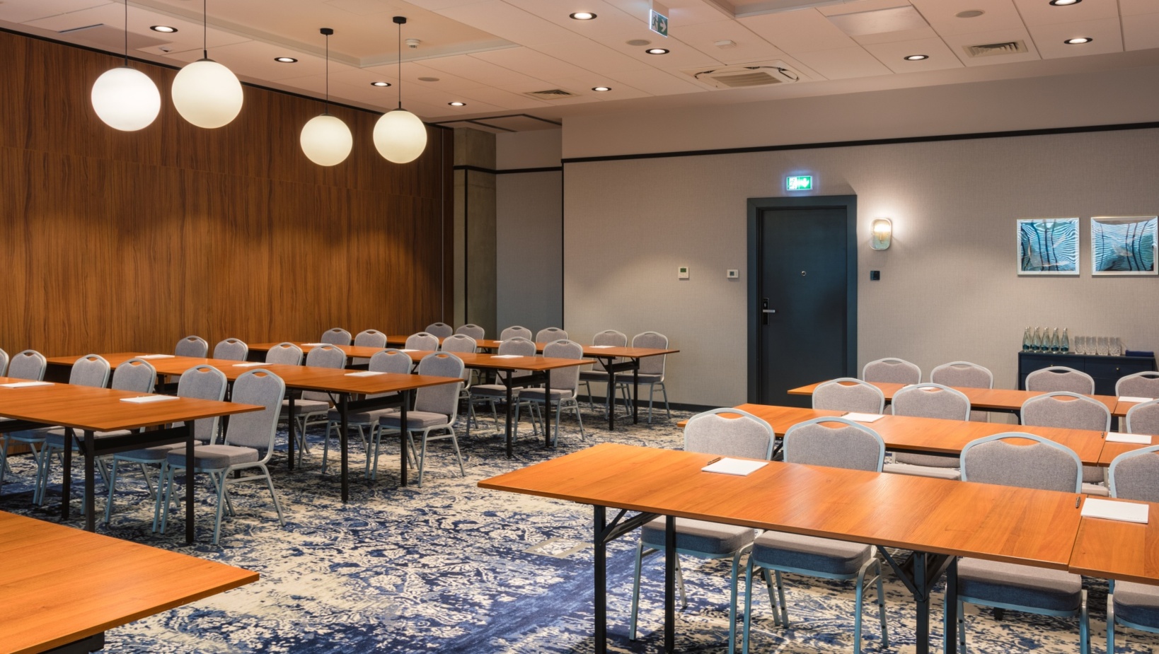Nowoczesna sala konferencyjna z drewnianymi stołami, szarymi krzesłami i dekoracyjnym dywanem, gotowa na szkolenie lub spotkanie.