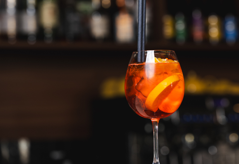 Glass of orange cocktail on bar counter garnished with orange slice and two black straws