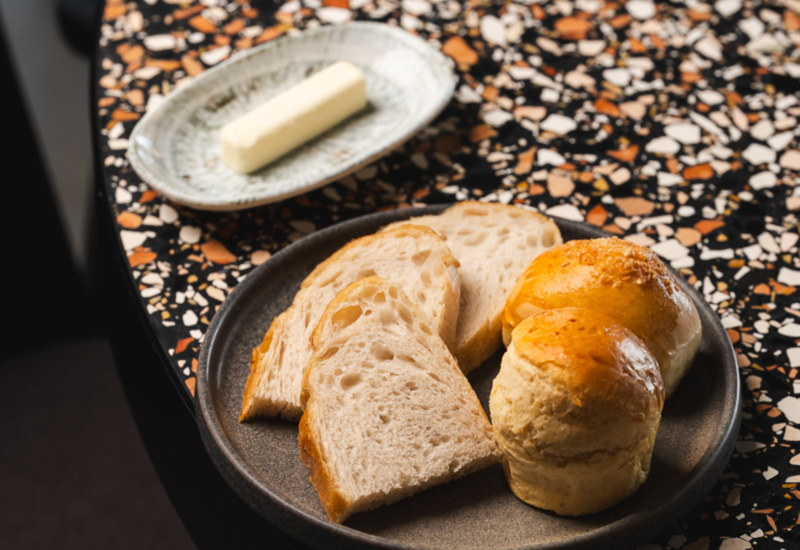 Plate with slices of fresh bread and two golden rolls on a terrazzo table, accompanied by a dish with a stick of butter