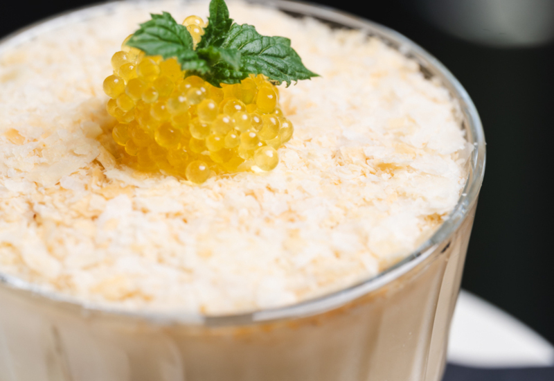 Creamy dessert in a glass topped with delicate flakes and yellow pearls garnished with a fresh mint leaf