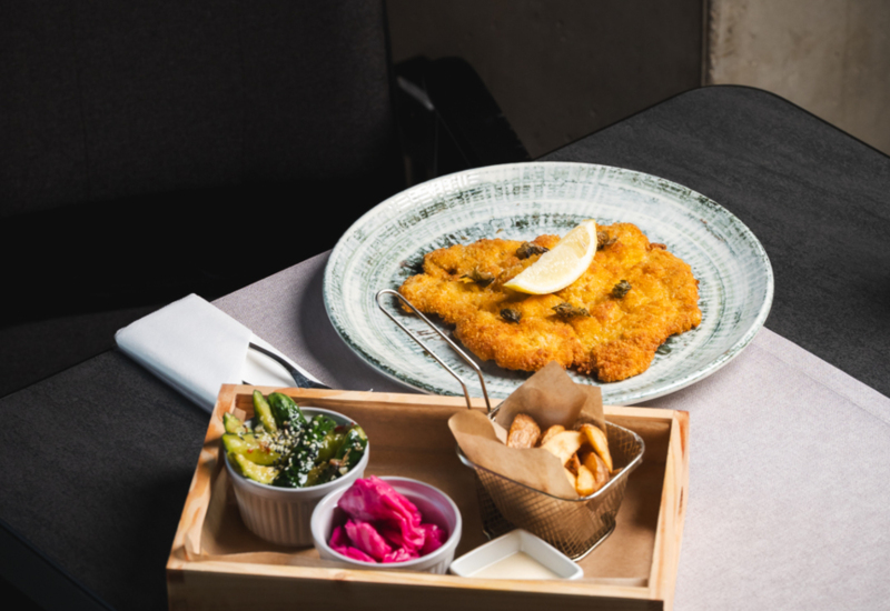 Crispy breaded pork cutlet with lemon wedge, served with fries, pickles, and salad on a wooden tray