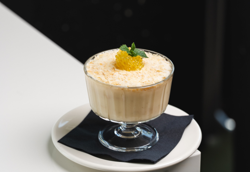 Creamy dessert served in a glass dish on a white plate with a black napkin, topped with yellow pearls and a mint leaf.