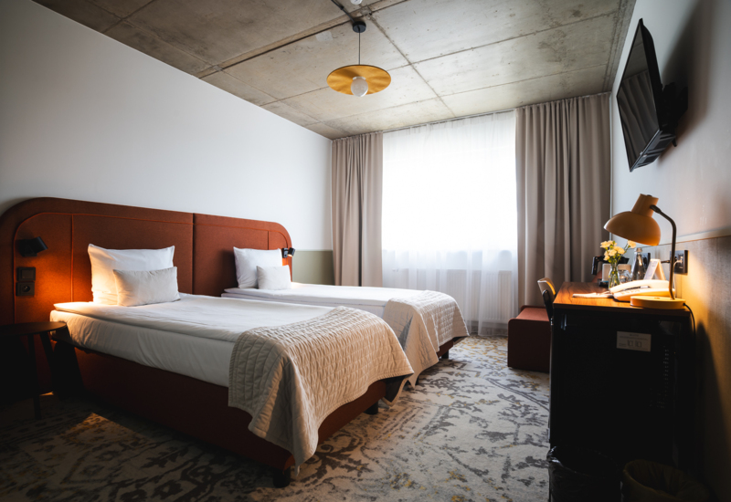 Hotel room with two beds, large window with curtains, desk with lamp, and wall-mounted TV, featuring modern decor.