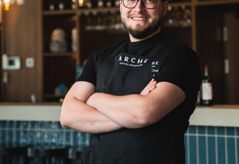 Smiling chef in a black apron stands in hotel restaurant with wooden shelves and bar stools in the background.