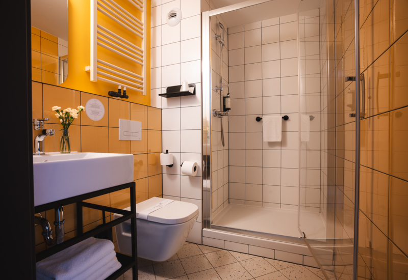 Modern hotel bathroom with shower, white fixtures, glass door, yellow tiles, and neatly arranged amenities