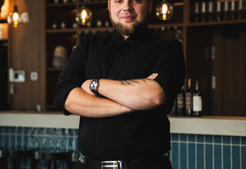 Smiling bar staff with crossed arms standing by a modern bar featuring blue tiles and hanging lights.
