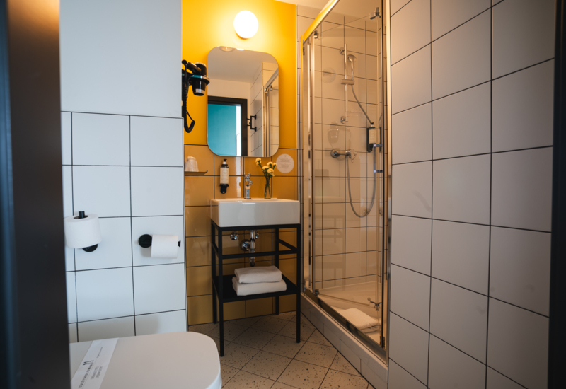 Modern hotel bathroom with black metal sink stand, glass door shower, and yellow accent wall behind the mirror