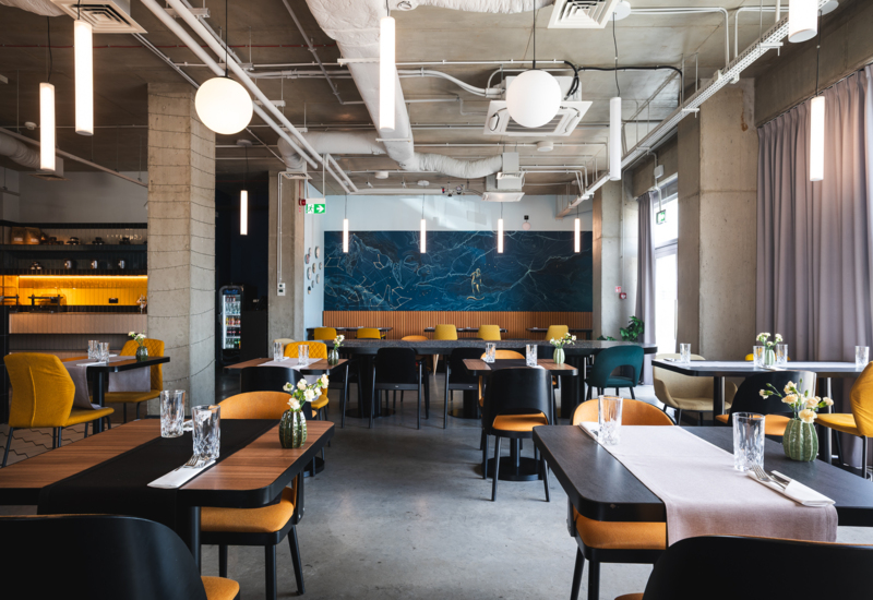 Modern hotel restaurant with industrial ceiling, wooden tables, yellow and black chairs, and small floral decorations on tables