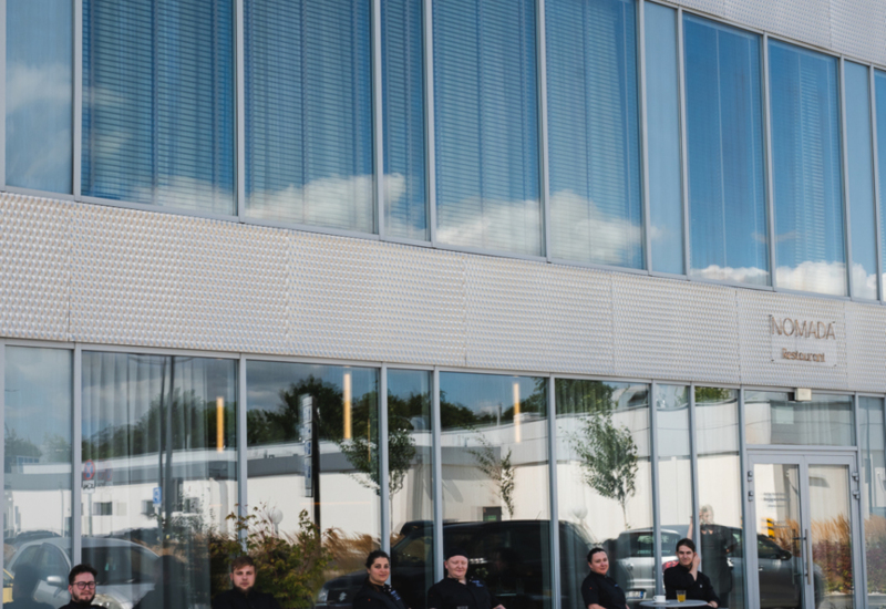 Staff members sit at outdoor tables in front of Nomada restaurant with large windows and modern metal facade.