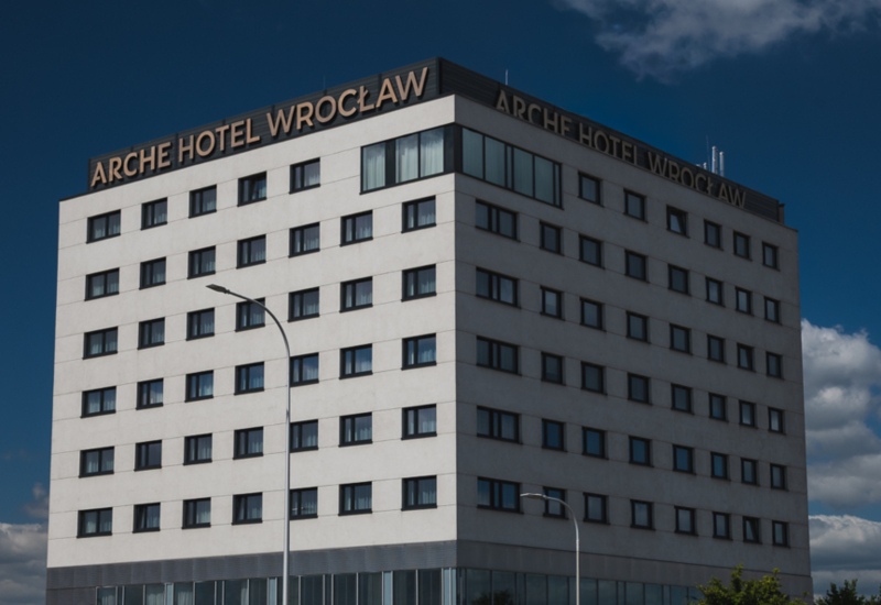 Modern nine-story Arche Hotel Wrocław with large windows and rooftop signage, located along a busy city street under a partly cloudy sky.