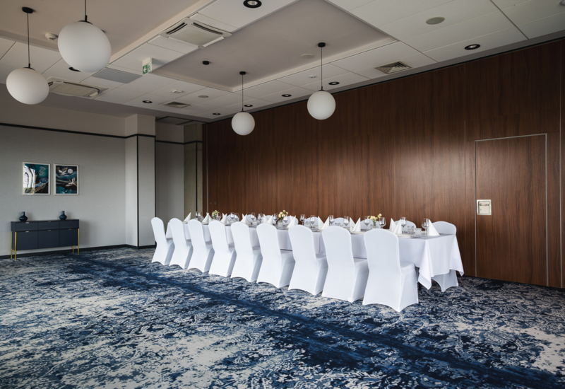 Elegant conference room with a large table covered in white cloth, white chair covers, wooden wall paneling, and modern pendant lighting.