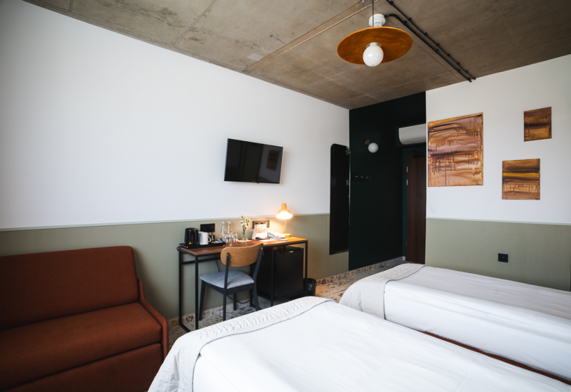 Modern hotel room with two single beds, a desk, TV, and a brown sofa in a minimalist setting.