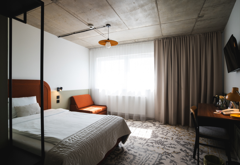 Modern hotel room with a large bed, orange sofa, wooden desk with lamp, and wall-mounted TV