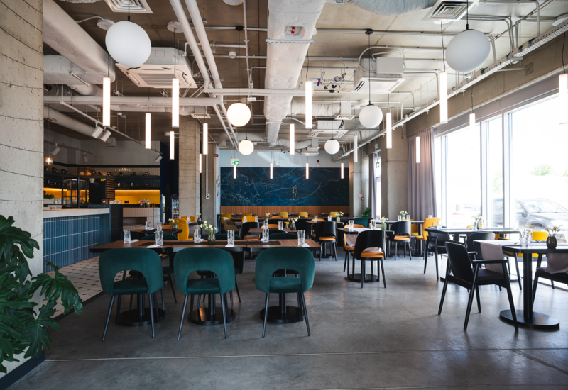Modern restaurant interior with comfortable green and yellow chairs, stylish lighting, and large windows letting in natural light.