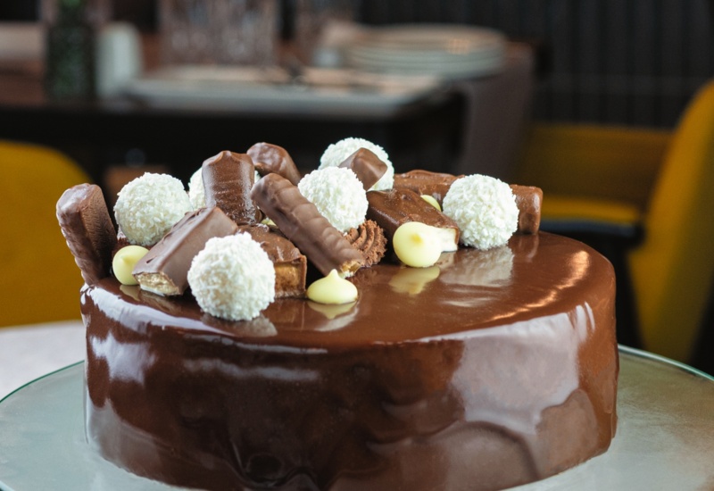 Glossy chocolate cake topped with assorted chocolate pralines and white coconut balls on a glass plate in a restaurant