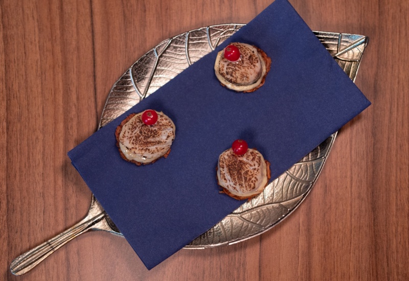 Three creamy mini desserts with toasted tops and a red berry on a silver leaf-shaped tray with a navy napkin
