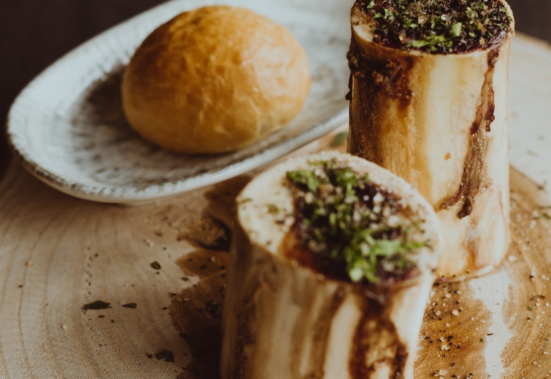 Roasted bone marrow garnished with herbs and spices on a wooden board, served with a fresh bread roll on the side.