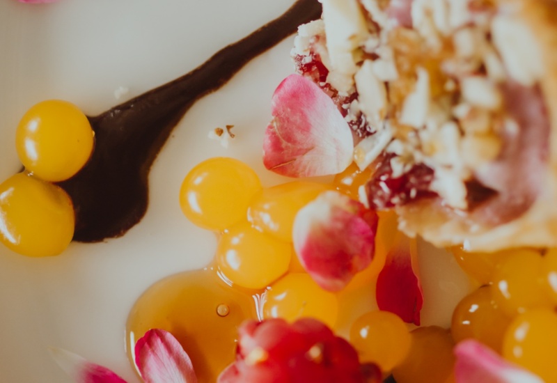 Artfully plated dessert with yellow fruit pearls, rose petals, and a chocolate sauce drizzle