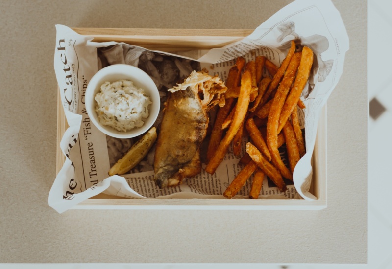 Battered fish with sweet potato fries and tartar sauce served on newspaper in a wooden tray