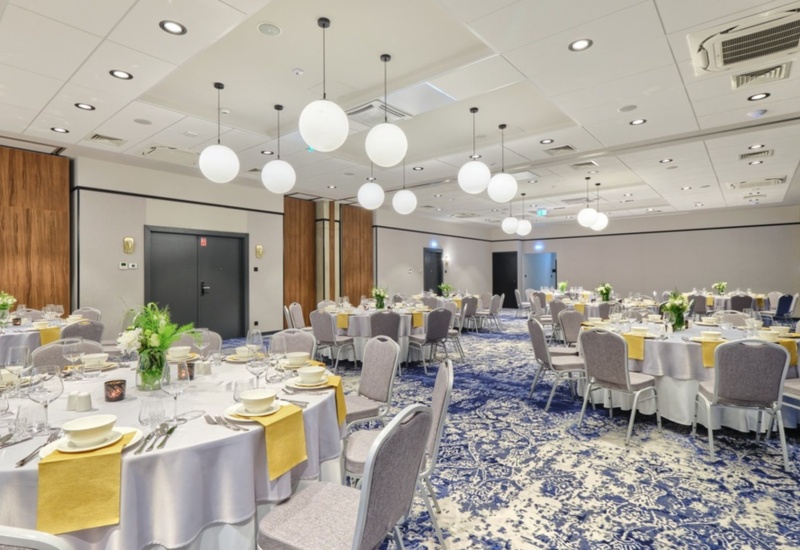 Elegant banquet hall with round tables draped in white linen and yellow napkins, gray chairs, and modern round pendant lights