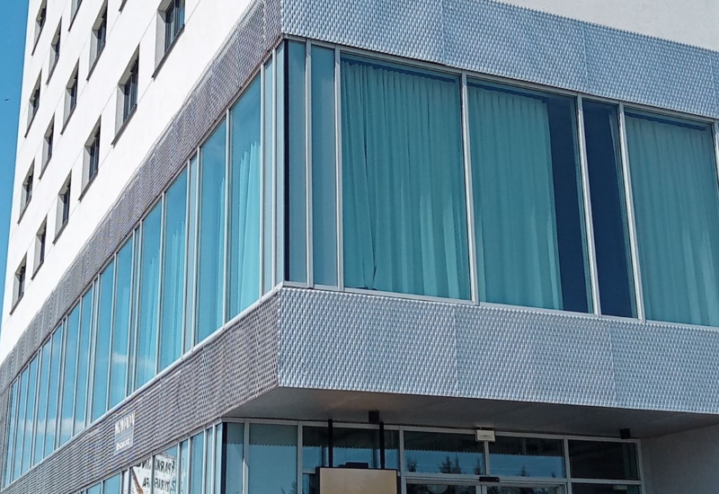 Modern hotel corner with large blue-curtained windows and silver metal mesh facade under clear blue sky