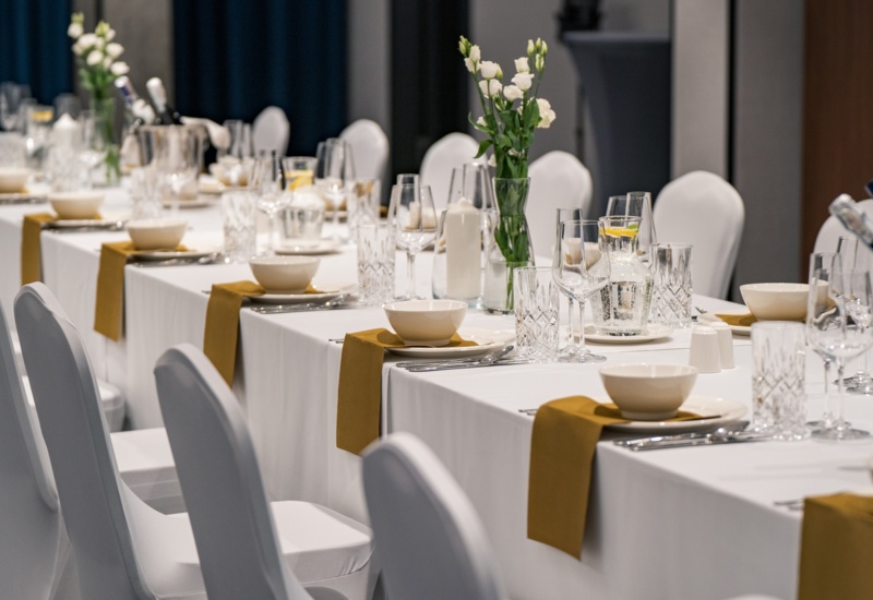 Long banquet table with white tablecloth, elegant glassware, gold napkins, and fresh flowers in vases set for event
