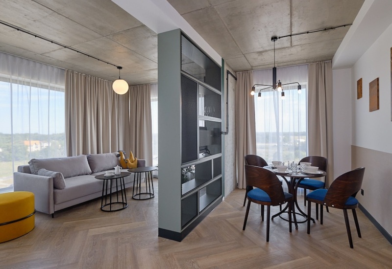 Modern hotel living area with large windows, gray sofa, yellow ottoman, dining table, and dark chairs with blue cushions.