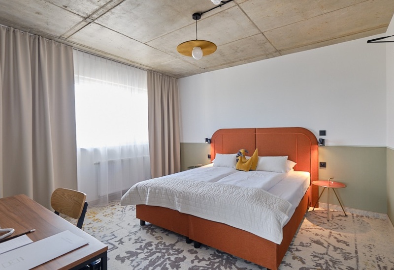 Modern hotel room with large orange double bed, light curtains, carpeted floor, and wooden desk with a lamp