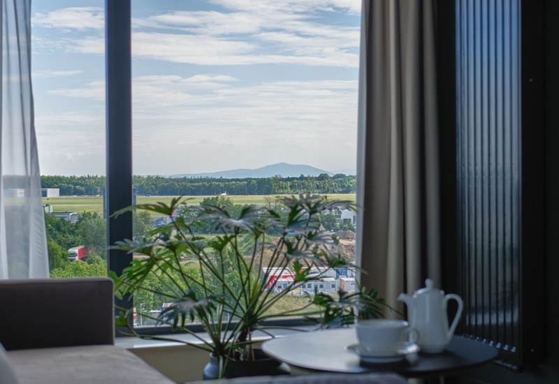 Hotel room with large window overlooking green landscape and distant hills, cozy sofa, and table with coffee set
