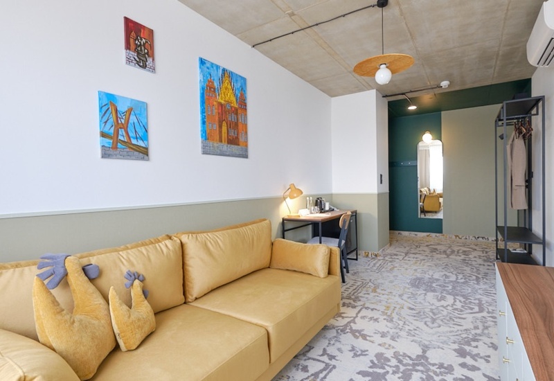 Modern hotel room with yellow sofa, desk, clothing rack, and colorful wall art in a bright, spacious setting