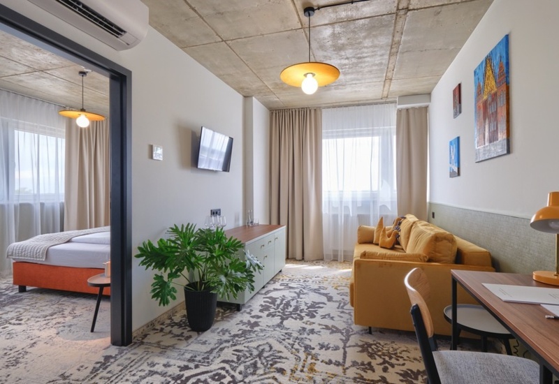 Modern hotel suite with a cozy sofa, work desk, decorative plant, and bedroom featuring a large bed and soft lighting.