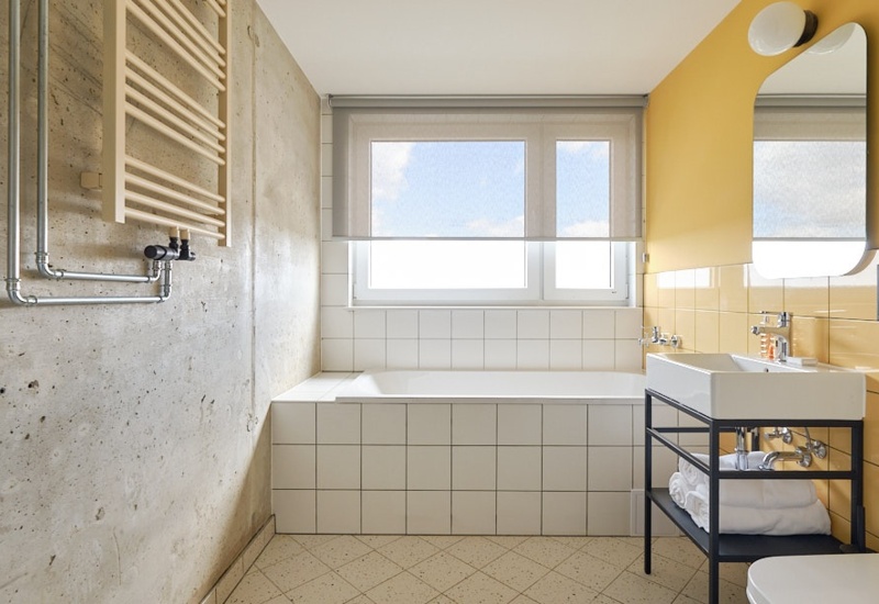 Hotel bathroom with a large built-in bathtub tiled in white, bright window, and yellow wall tiles creating a cheerful space.