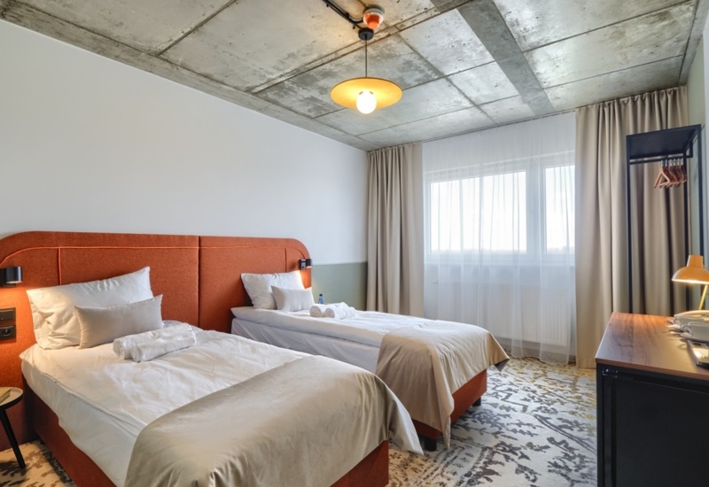 Hotel room with two single beds featuring orange headboards, desk, TV, and large window with curtains.