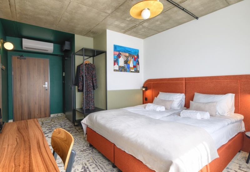 Modern hotel room with a large bed, orange headboard, minimalist wardrobe, and wooden desk with kettle and cups.
