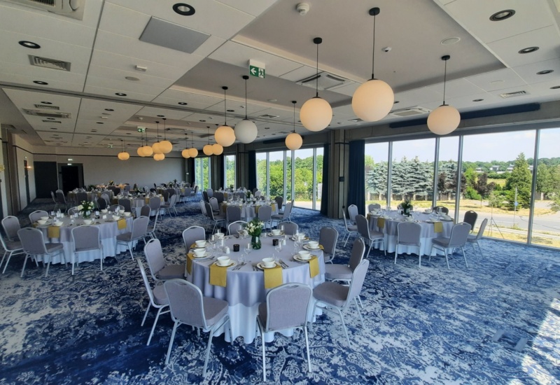 Spacious banquet hall with round tables, elegant decor, and large windows overlooking greenery outside.