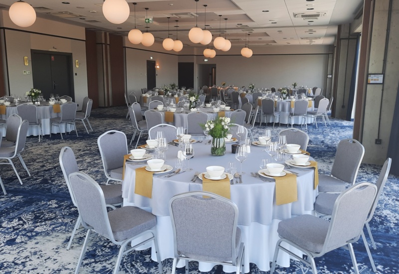 Spacious banquet hall with round tables, white linens, gray chairs, yellow napkins, and floral centerpieces, set for a formal event.