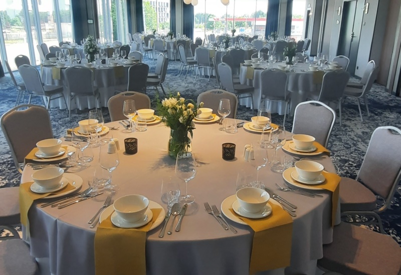 Banquet room with round tables set with white linens, yellow napkins, floral centerpieces, and natural light through large windows