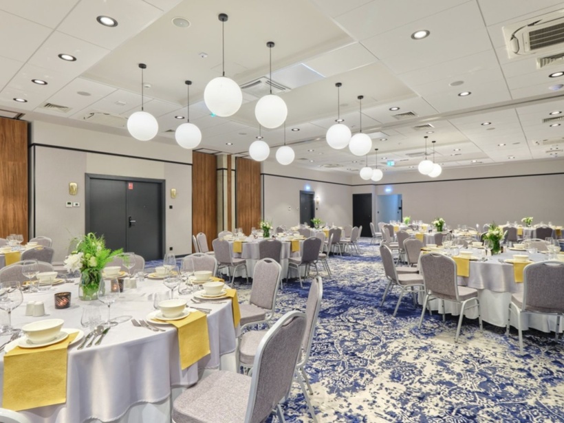 Elegancka sala bankietowa z okrągłymi stołami nakrytymi białymi obrusami i żółtymi serwetkami, szare krzesła, nowoczesne oświetlenie kuliste