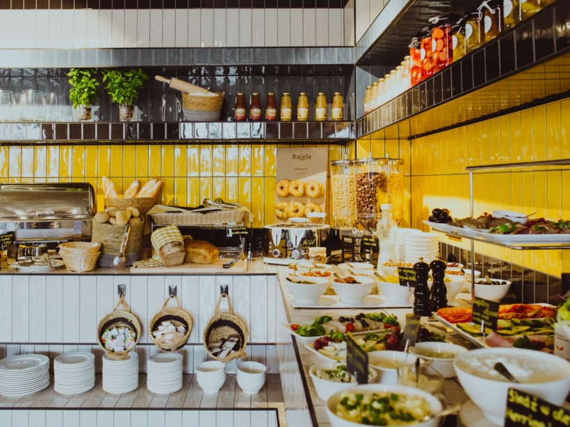 Bufet śniadaniowy z różnorodnym pieczywem, płatkami, świeżymi sałatkami i sokami w jasnej, żółtej kuchni.