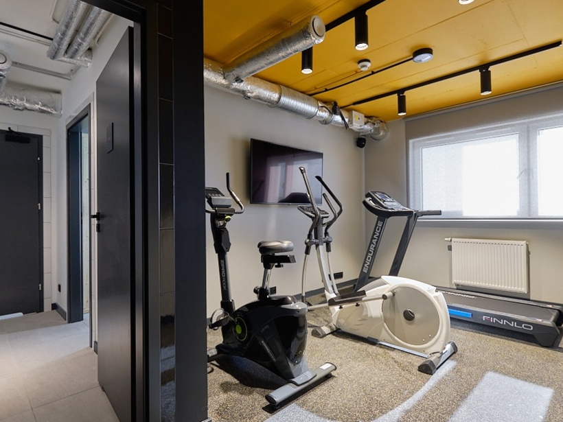 Mała siłownia hotelowa z rowerem stacjonarnym, orbitrekiem, bieżnią i dumbbellami, jasne wnętrze z oknem i żółtym sufitem.